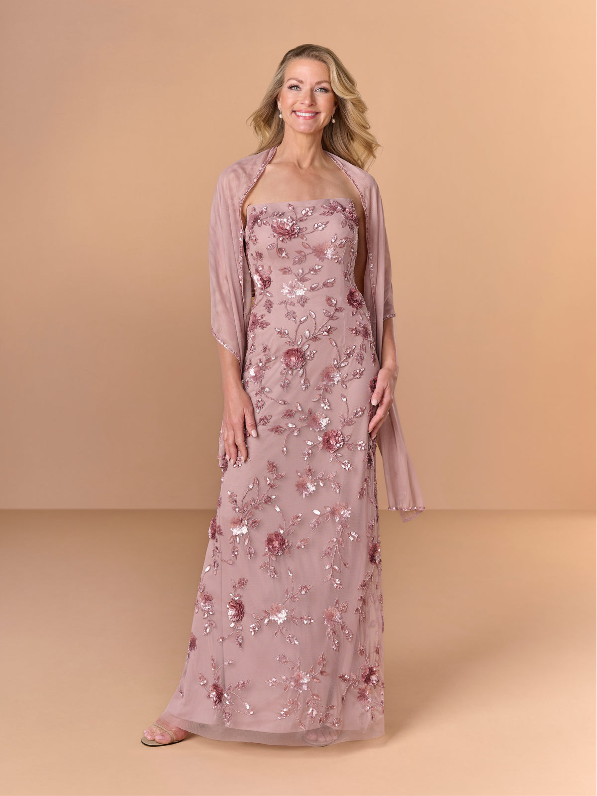 A woman models a floor-length dusty rose formal gown with floral sequin embroidery, paired with a matching sheer shrug, against a solid beige studio backdrop. / All over beaded gown with high straight neckline, 3D flowers and matching separate shawl.