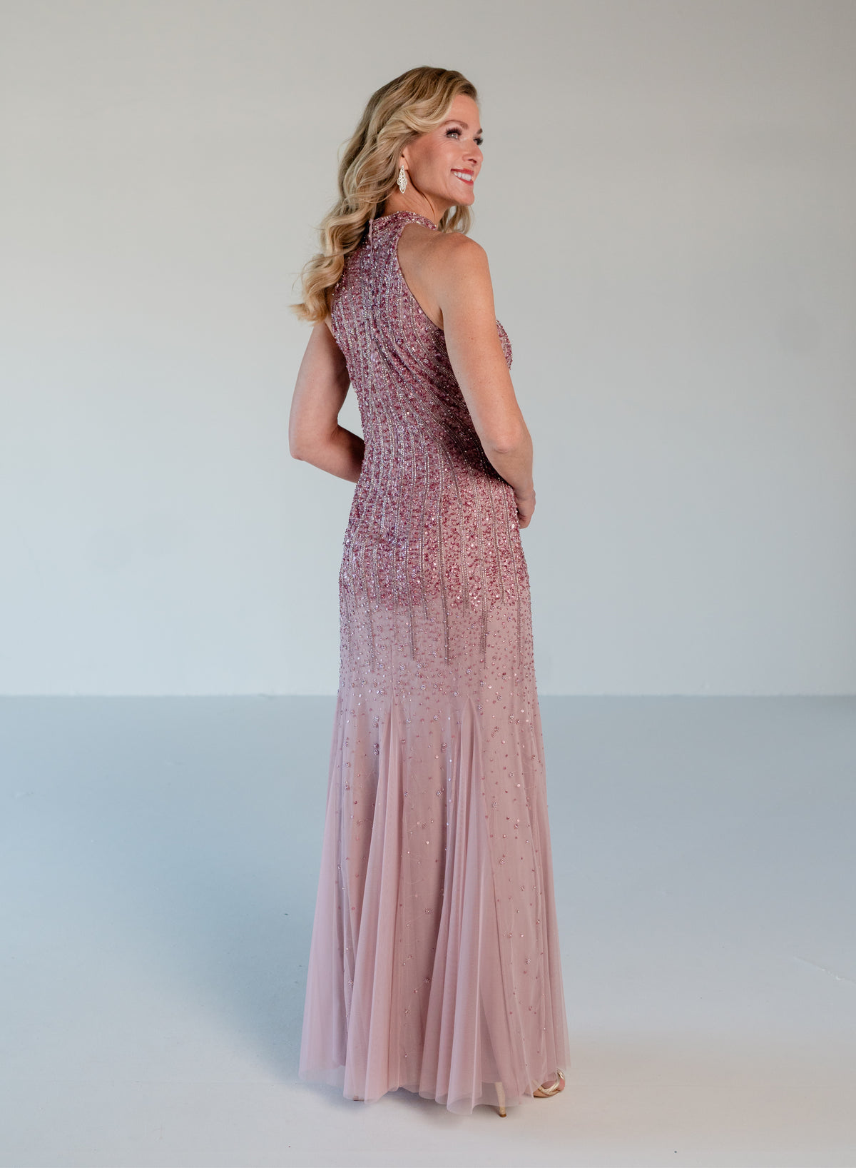 A woman modeling a sleeveless, high-neck, floor-length gown with dense beaded embellishment and a flowing tulle skirt, photographed against a plain light gray studio background. / Halter neckline slim gown with soft godets and all over ombre beading.