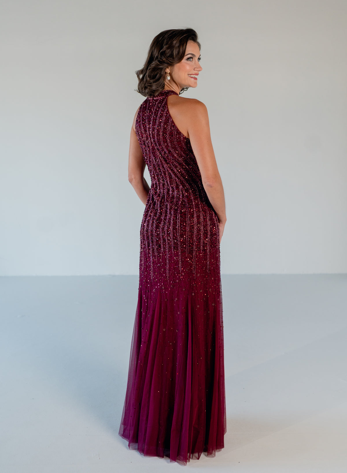 A woman modeling a sleeveless, high-neck floor-length gown with a fitted bodice covered in vertical beaded stripes and a flowing tulle skirt, shown from the back against a plain light gray studio background. / Halter neckline slim gown with soft godets and all over ombre beading.