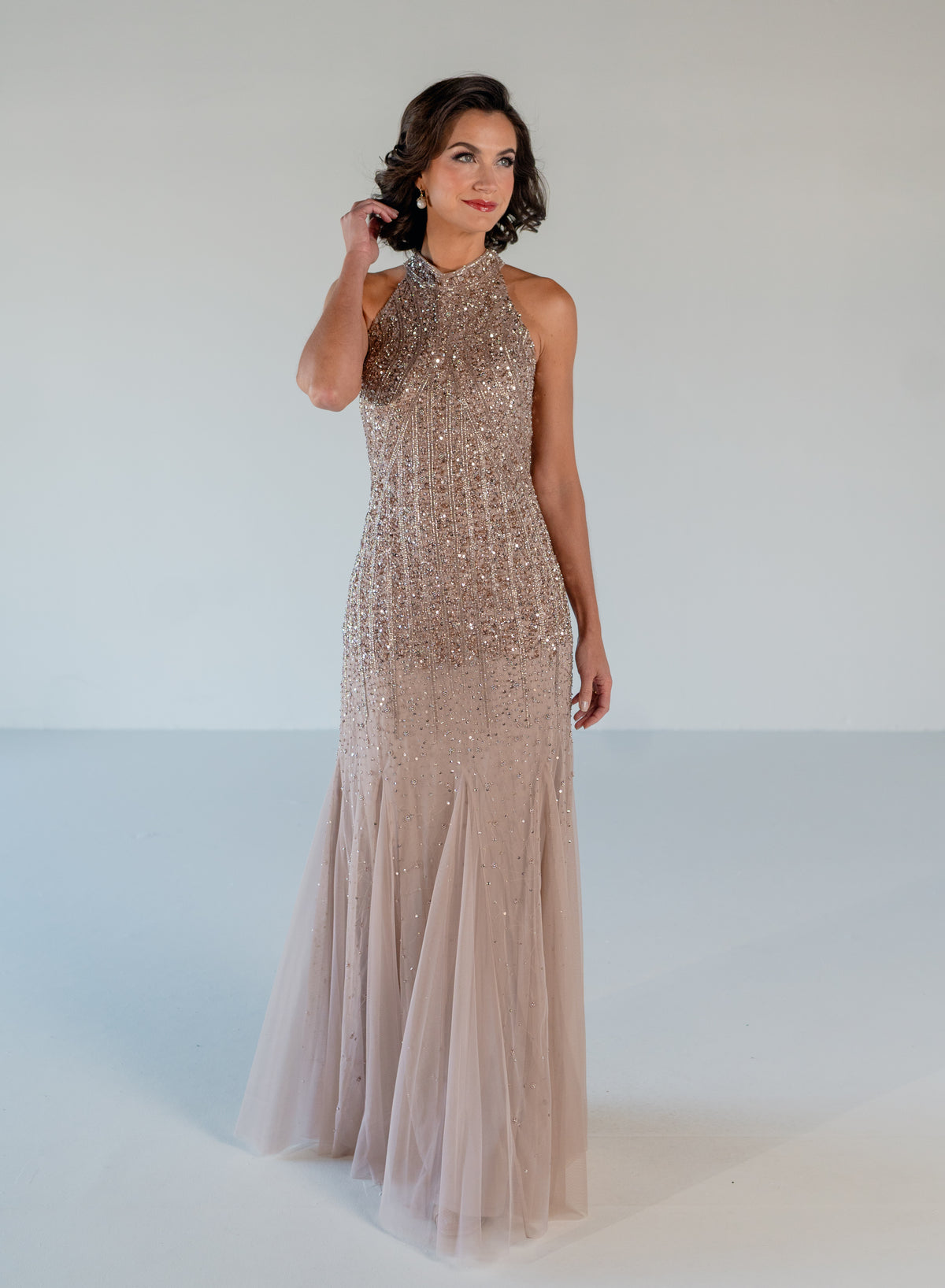 A woman modeling a sleeveless, high-neck floor-length gown covered in shimmering sequins and beadwork, with a flowing tulle skirt, against a plain light gray studio background. / Halter neckline slim gown with soft godets and all over ombre beading.