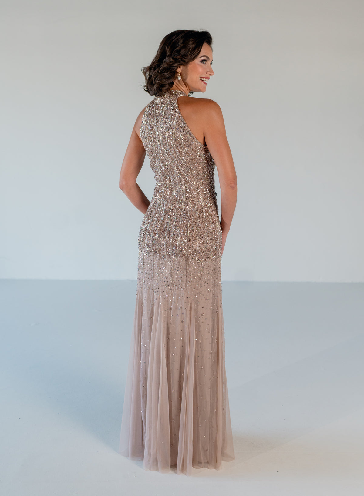 A woman modeling a sleeveless, one-shoulder floor-length gown with dense beadwork and tulle, shown from the back against a plain light gray studio background. / Halter neckline slim gown with soft godets and all over ombre beading.