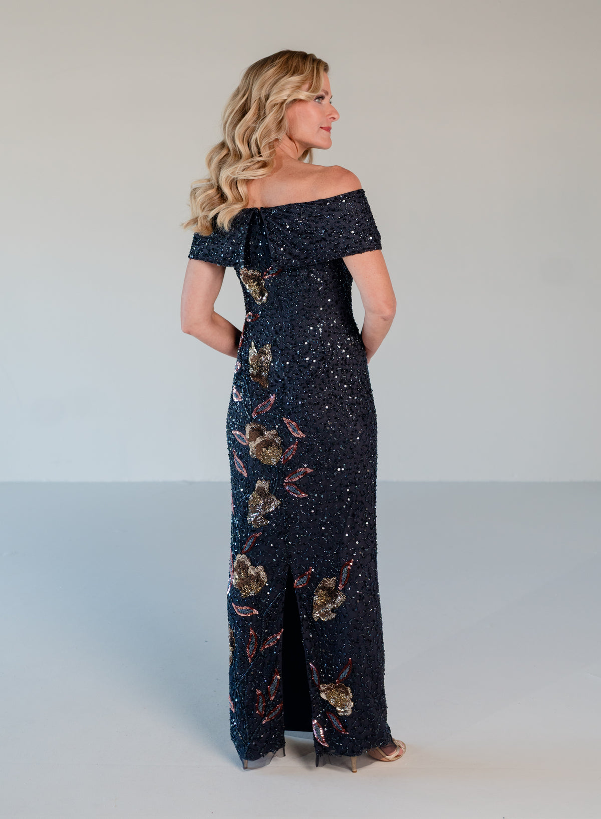 A woman modeling an off-the-shoulder, floor-length navy blue sequined gown with gold and pink floral embellishments, shown from a side-back angle against a light gray studio background. / Off the shoulder column gown with all over beading and multi color detailing.