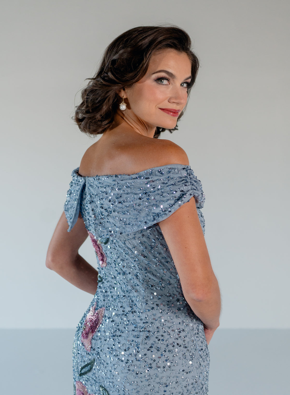 A woman wearing a light blue sequined off-the-shoulder evening gown, photographed from the back-three-quarter angle against a plain light gray studio background. / Off the shoulder column gown with all over beading and multi color detailing.