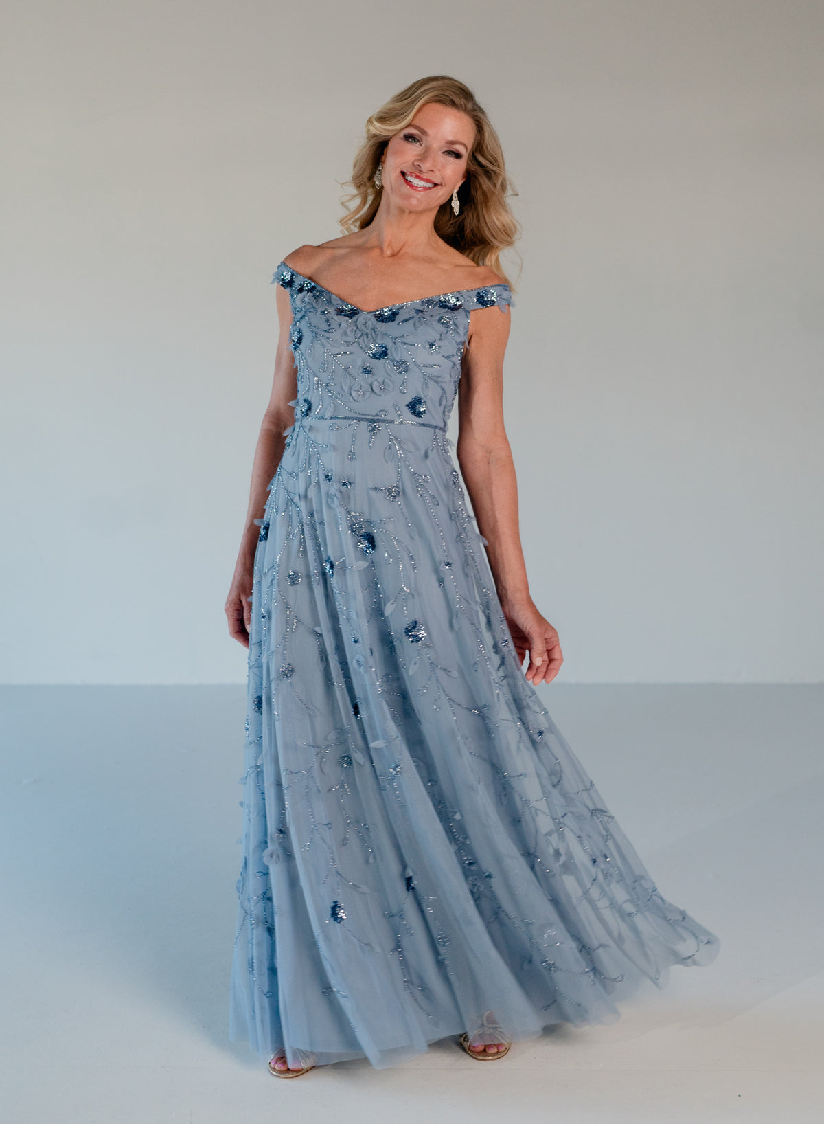 A woman modeling an off-the-shoulder light blue tulle maxi dress with intricate floral embroidery and beadwork, set against a plain light gray studio background. / Tip of the shoulder, soft A-line gown with 3D floral beading.