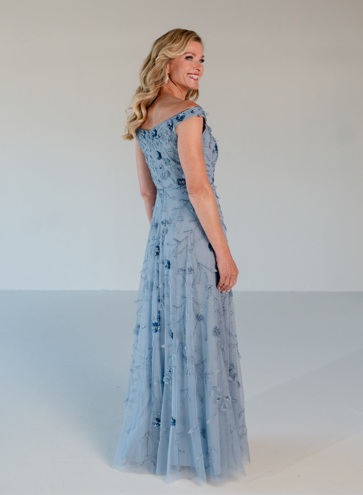 A woman modeling a floor-length light blue off-the-shoulder gown with a fitted bodice and flowing tulle skirt, photographed against a plain light gray studio background. / Tip of the shoulder, soft A-line gown with 3D floral beading.