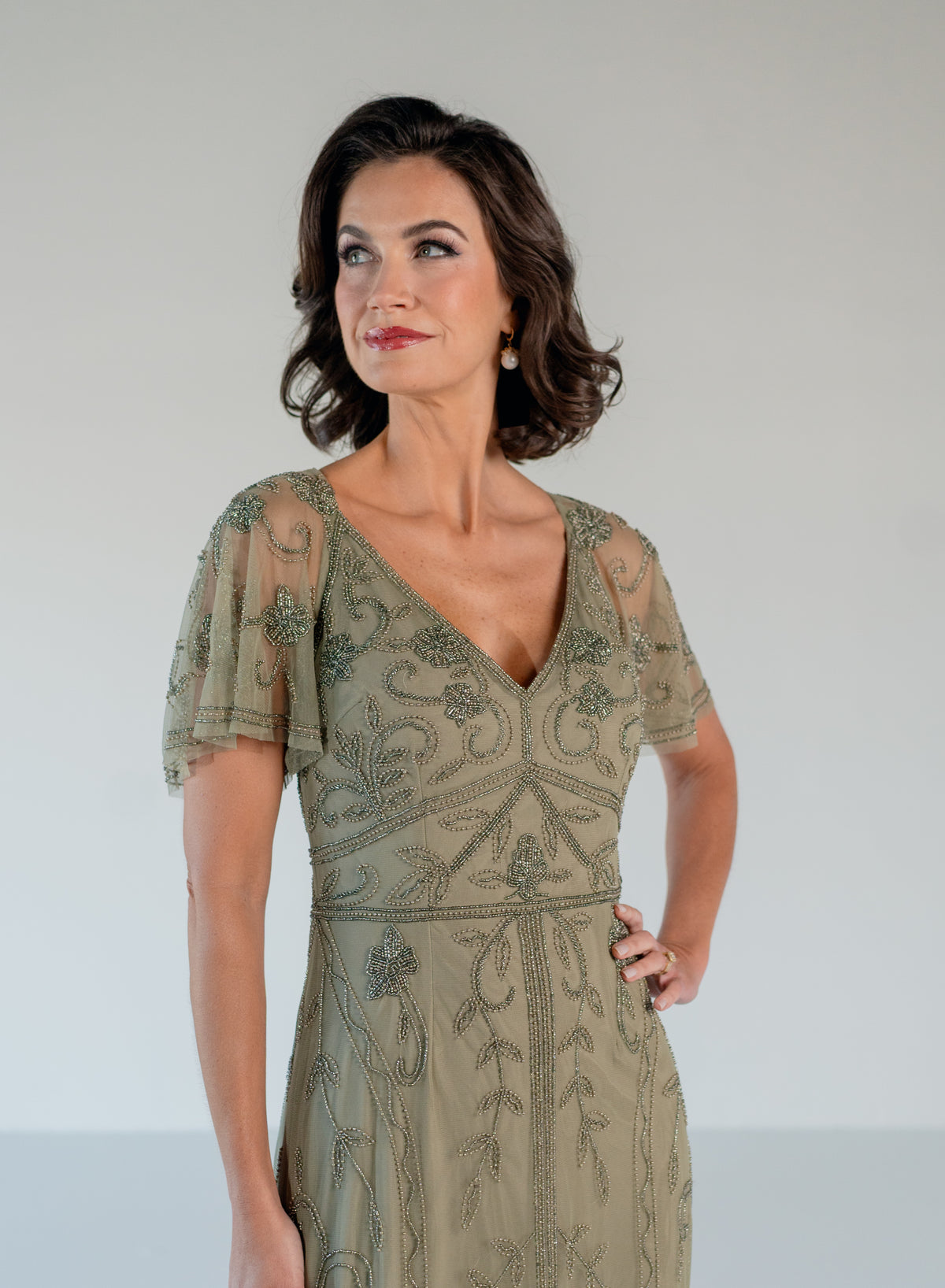 A woman modeling a sage green embroidered midi dress with sheer flutter sleeves and a V-neckline, set against a plain light gray studio background. / V neckline, soft flutter sleeve gown with godet skirt and all over beading.
