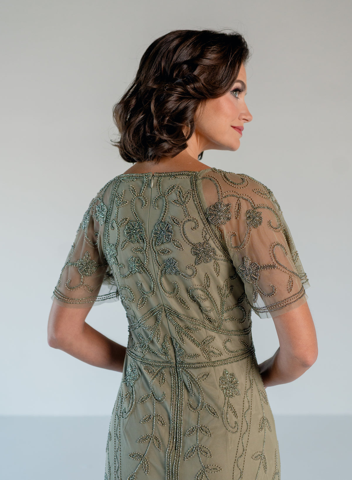 A woman shown from the back wearing a short-sleeved sage green dress with intricate floral embroidery and beadwork, photographed against a neutral light gray studio background. / V neckline, soft flutter sleeve gown with godet skirt and all over beading.