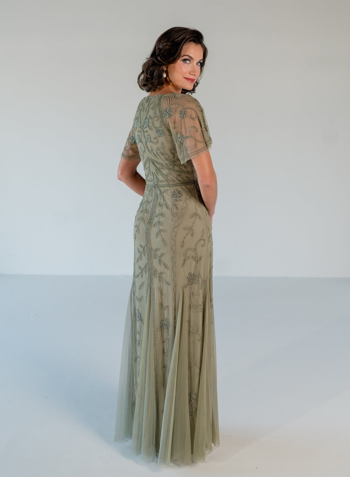 A woman modeling a floor-length sage green evening gown with sheer flutter sleeves and delicate floral embroidery, photographed against a plain light gray studio background. / V neckline, soft flutter sleeve gown with godet skirt and all over beading.
