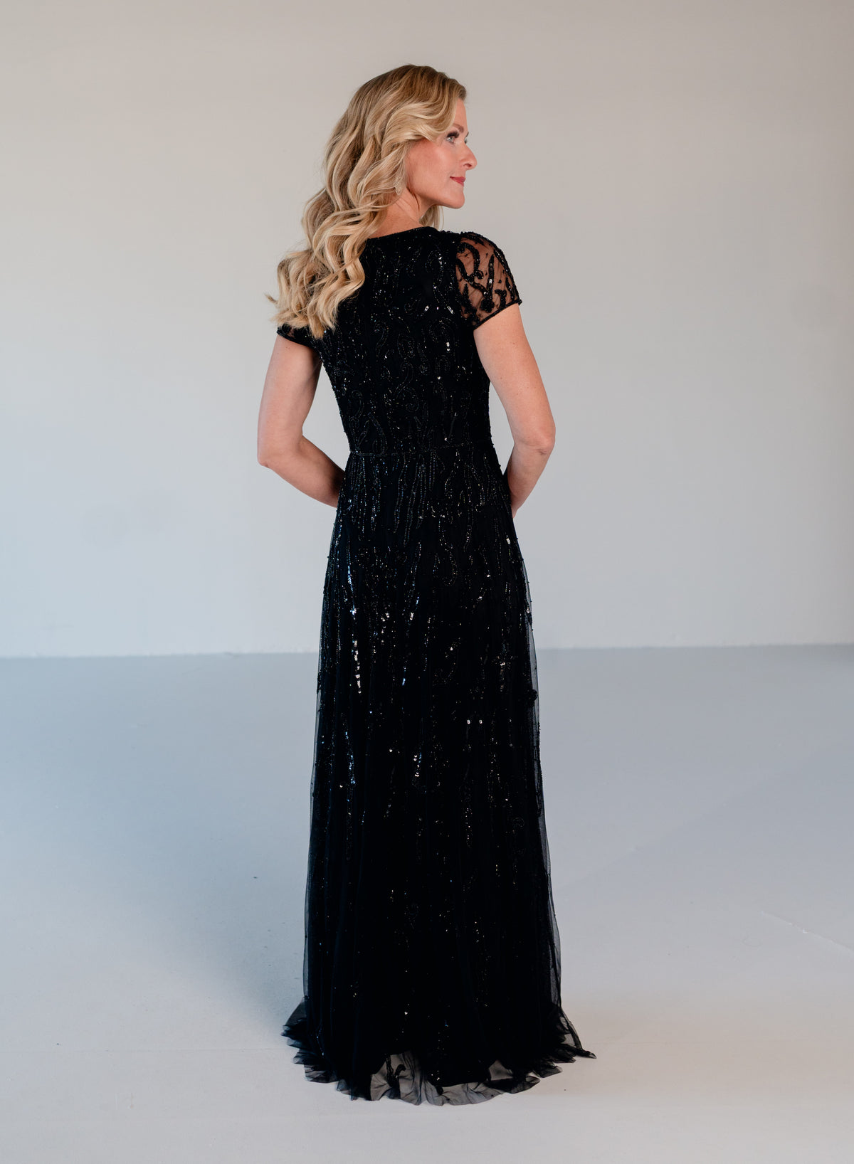 A woman modeling a floor-length black evening gown with lace short sleeves and a sequined tulle skirt, photographed from the back against a plain light gray studio backdrop. / Sweetheart neckline A-line gown with all over beading.