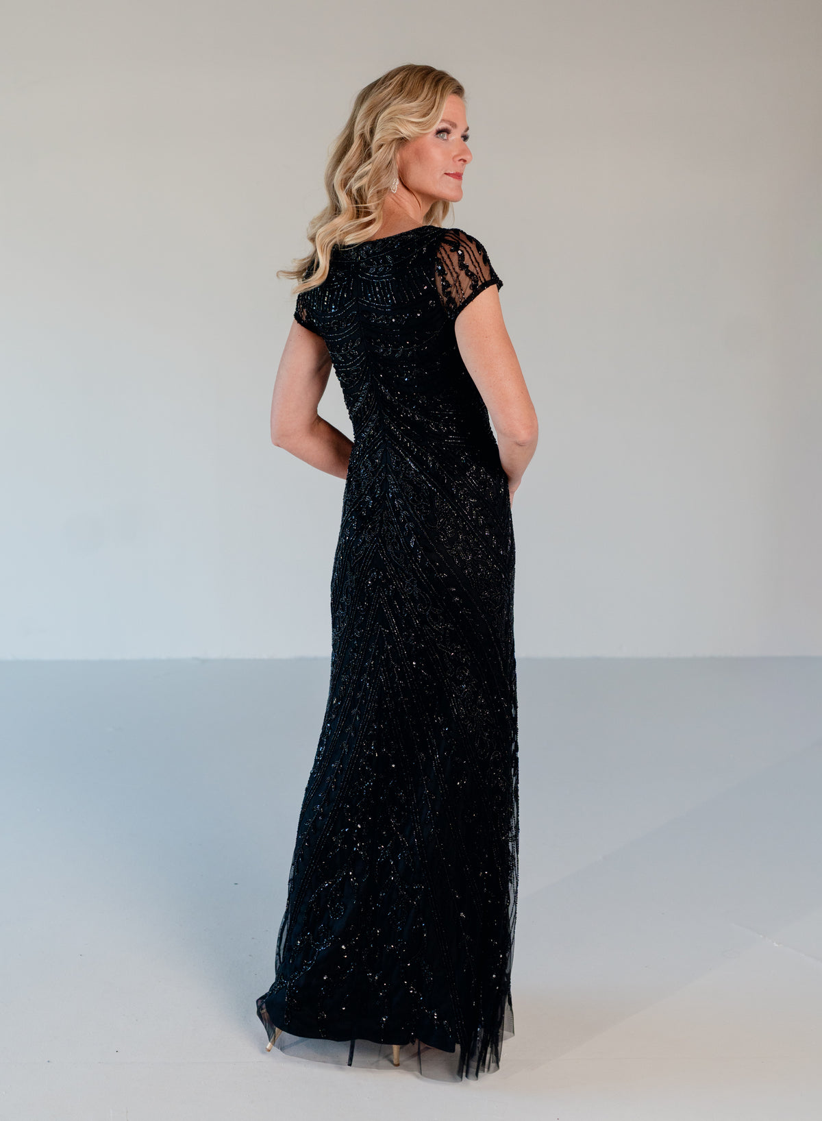 A woman modeling a floor-length black evening gown with cap sleeves and a sheer lace upper back, shown from the side against a plain light gray studio background. / Scoop neckline, slim gown with all over beading.