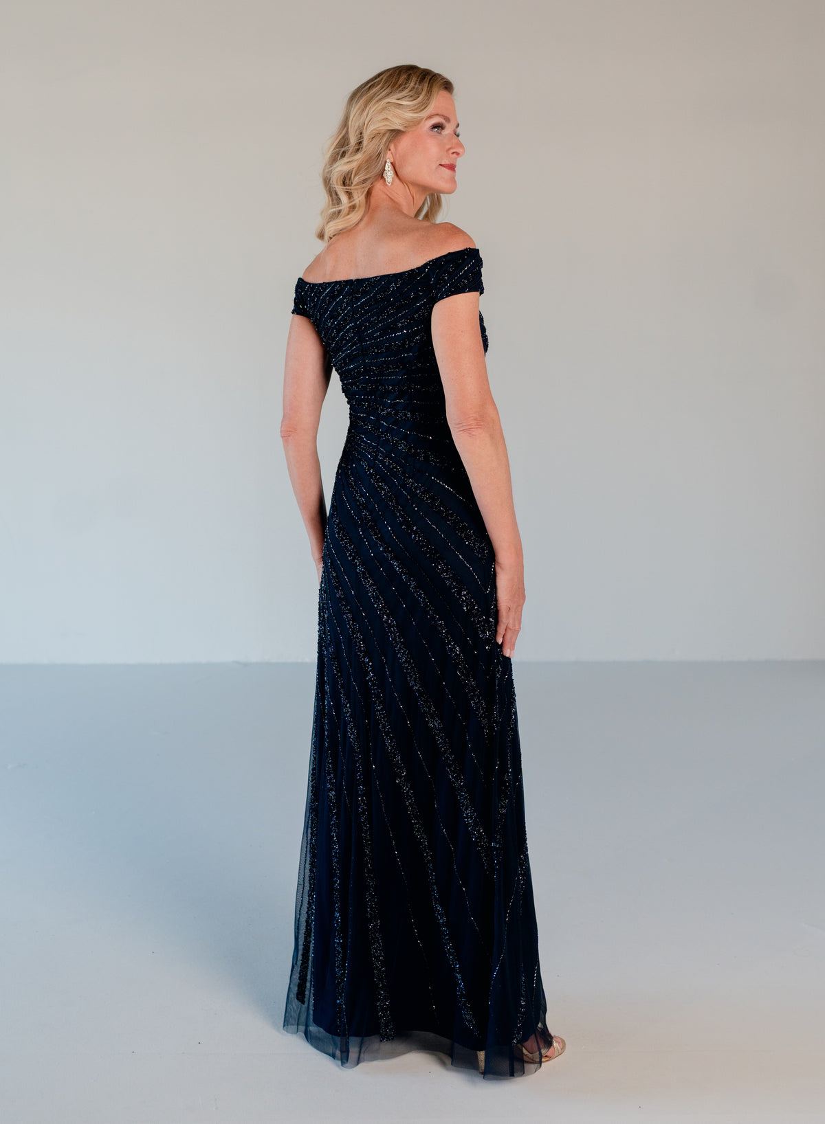 A woman modeling an off-the-shoulder, floor-length navy evening gown with subtle shimmering vertical stripes, photographed in a studio setting against a light gray background. / Off the shoulder slim gown with side rushing and linear beading.
