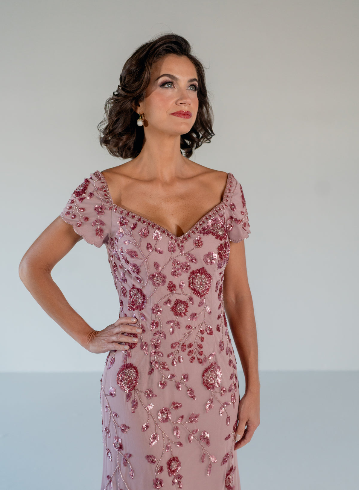 A woman modeling a dusty pink evening gown with rose gold sequin floral embroidery and short flutter sleeves against a plain light gray background. / Sweetheart neckline, slim gown with all over floral beading.