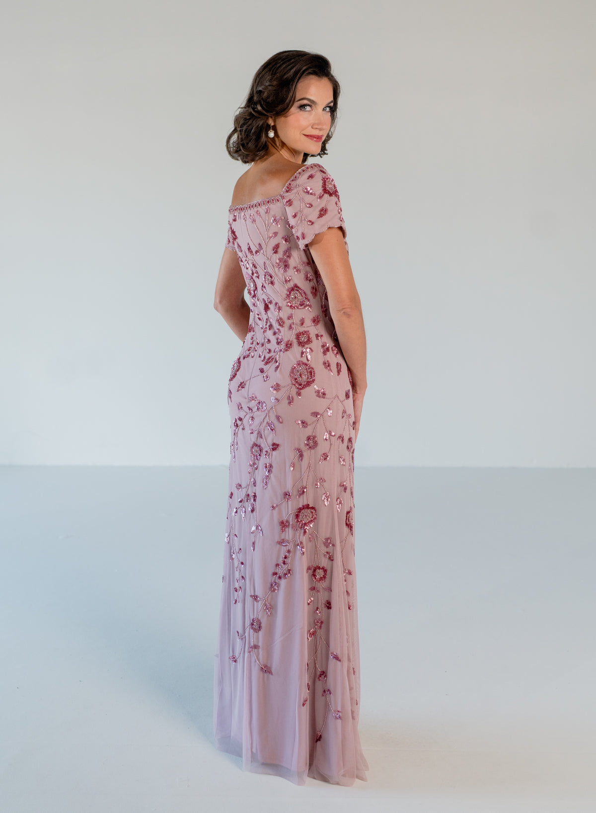 A woman modeling a floor-length dusty pink gown with short puff sleeves and an allover rose floral sequin and bead embellishment, photographed against a seamless light gray studio background. / Sweetheart neckline, slim gown with all over floral beading.