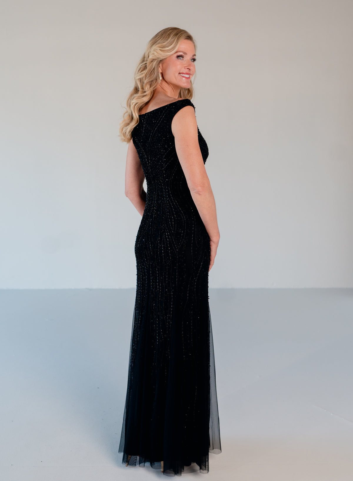 A woman modeling a sleeveless black evening gown with a fitted bodice, beaded texture, and sheer tulle overskirt, photographed against a plain light gray studio background. / Off the shoulder slim gown with soft godets and all-over linear beading.