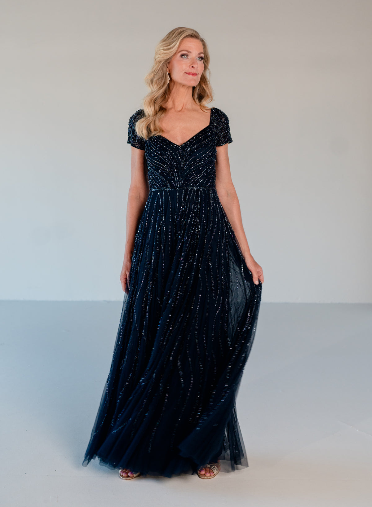 A woman modeling a floor-length navy blue evening gown with short cap sleeves, a sweetheart neckline, and shimmering vertical beadwork and tulle detailing, photographed against a plain light gray studio background. / Sweetheart neckline-line gown with all over trailing beading.