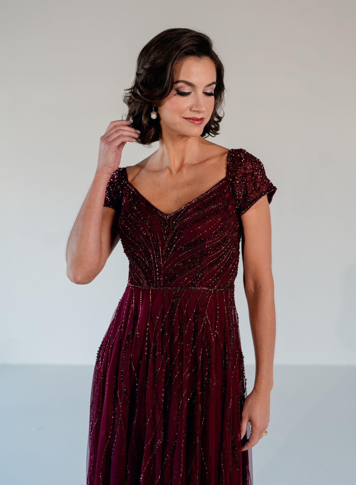 A woman wearing a floor-length burgundy evening gown with sparkling beadwork and cap sleeves, photographed against a plain light gray studio background. / Sweetheart neckline-line gown with all over trailing beading.