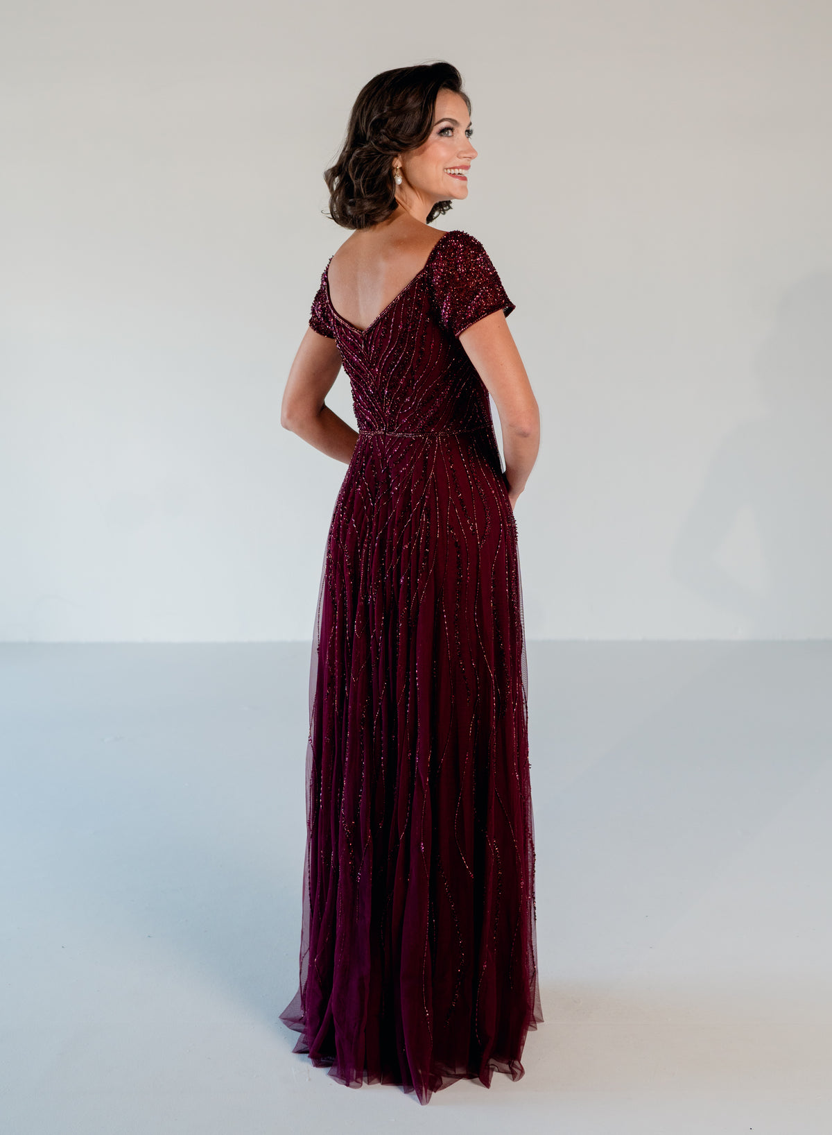 A woman modeling a floor-length burgundy evening gown with short cap sleeves, a deep V-neckline, and shimmering beaded detailing, photographed against a plain light gray studio background. / Sweetheart neckline-line gown with all over trailing beading.