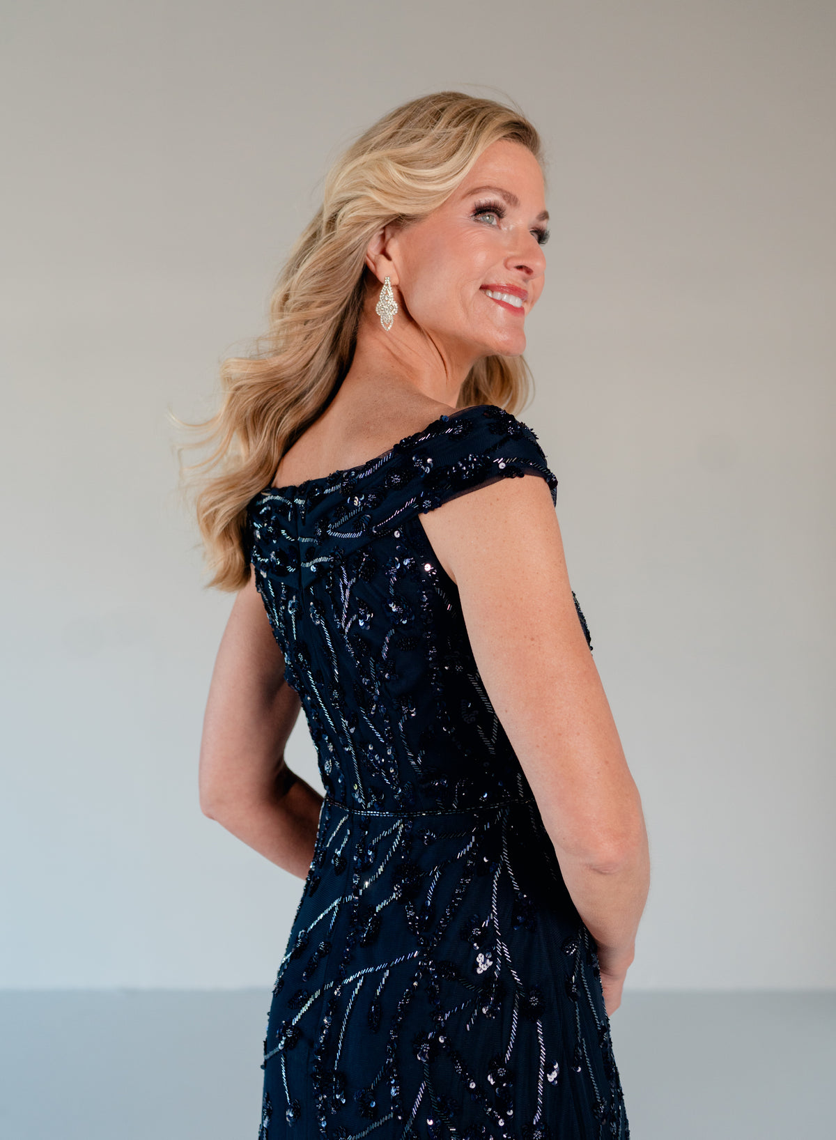 A blonde woman wearing a dark navy off-the-shoulder evening gown with shimmering sequin details, photographed against a plain light gray studio background. / Off the shoulder A-line gown with all over floral and trailing beading.