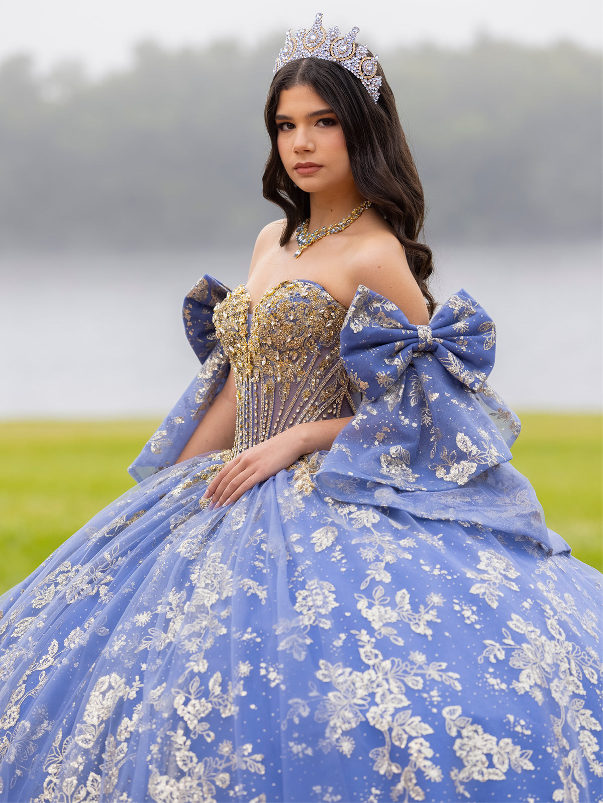 Woman in a blue and gold formal gown with a large bow and tiara outdoors. / This ball gown features an embellished bodice and off-the-shoulder sleeves, paired with a full skirt decorated with intricate floral detailing and a large back bow.