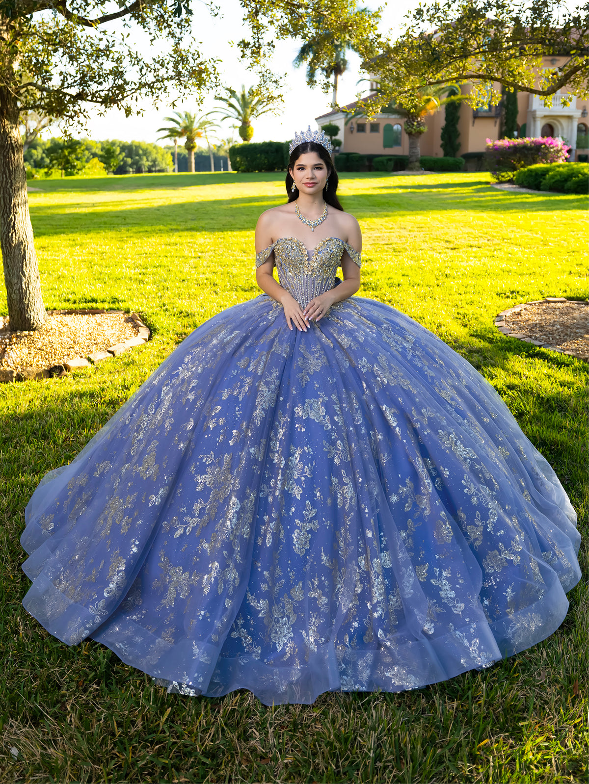 A young woman wearing a voluminous lavender-blue ball gown with silver floral embroidery and a sparkly embellished bodice, standing outdoors on a sunny grassy lawn with trees and a house in the background. / This ball gown features an embellished bodice and off-the-shoulder sleeves, paired with a full skirt decorated with intricate floral detailing and a large back bow.