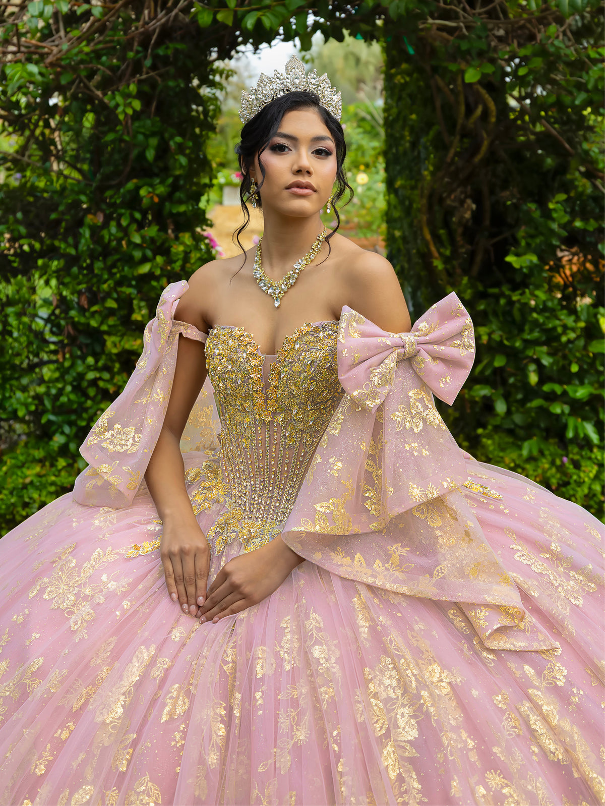 A woman wearing an elaborate pink and gold ball gown with a tiara, posed outdoors in front of a lush green foliage background. / This ball gown features an embellished bodice and off-the-shoulder sleeves, paired with a full skirt decorated with intricate floral detailing and a large back bow.