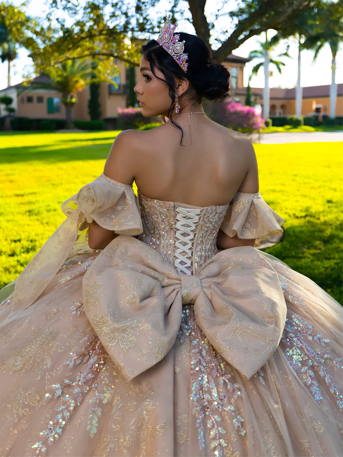 A woman photographed from behind wearing an elaborate off-the-shoulder ball gown with a large bow at the back, sequined tulle, and a pink crown, standing outdoors in a sunny grassy yard with houses and trees in the background. / This ball gown features an embellished bodice, off-the-shoulder sleeves, and a full skirt with floral detailing. A large bow at the back completes the look.