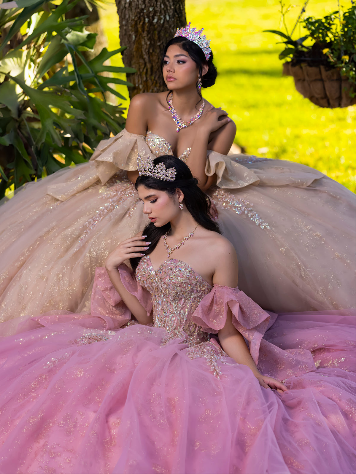 Two young women in elaborate ball gowns pose outdoors in a garden. The seated woman in the foreground wears a pink tulle quinceañera-style gown with gold embellishment and a tiara, while the standing woman behind her wears a champagne/beige gown with similar detailing and a tiara. / This ball gown features an embellished bodice, off-the-shoulder sleeves, and a full skirt with floral detailing. A large bow at the back completes the look.