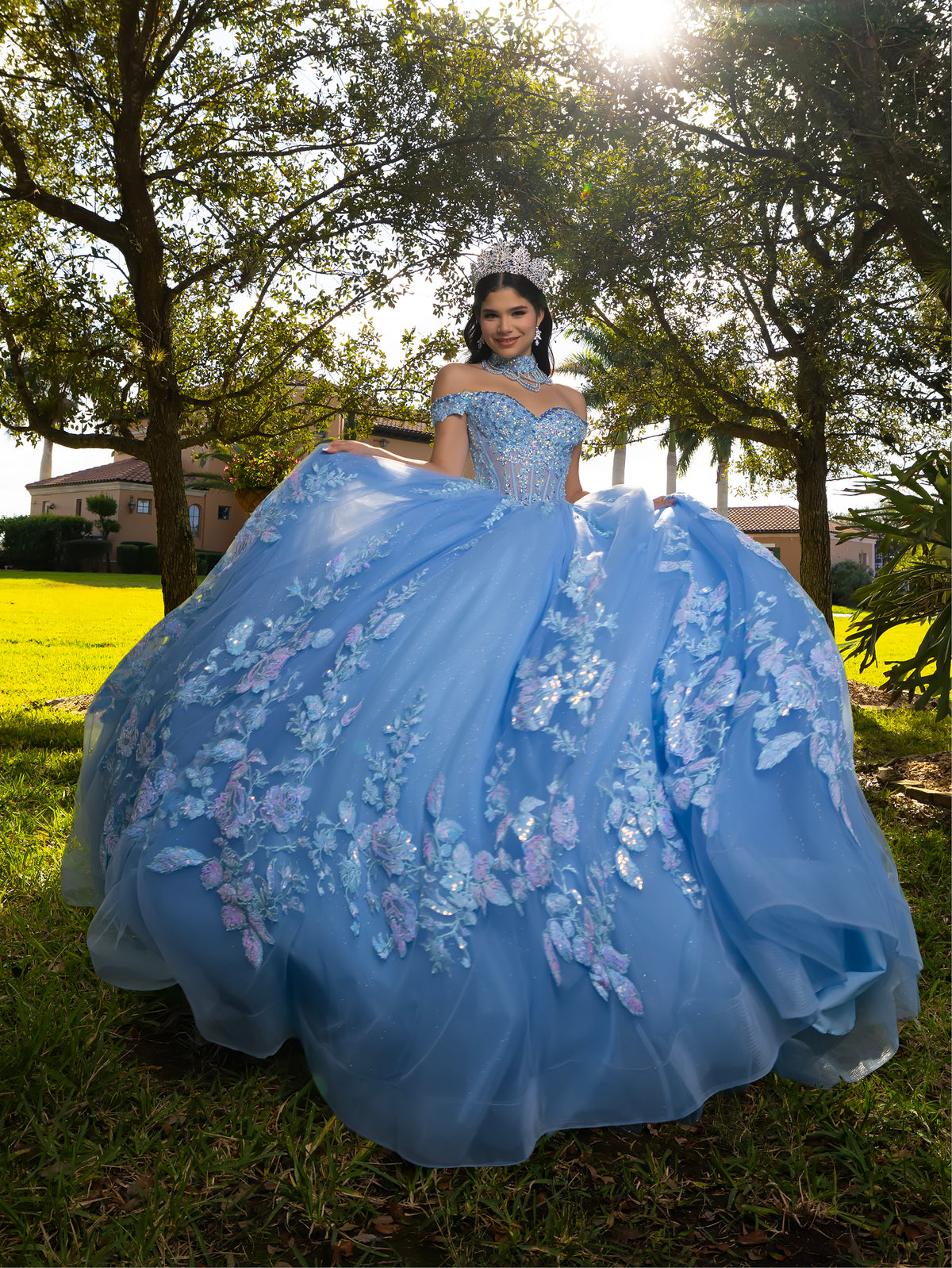 A woman wearing a voluminous light blue quinceañera ball gown with floral lace appliqués, standing outdoors in a grassy park with trees and sunlight in the background. / This ball gown features an embellished bodice, off-the-shoulder sleeves, and a full skirt covered in detailed floral appliqué. The back is finished with a large decorative bow.
