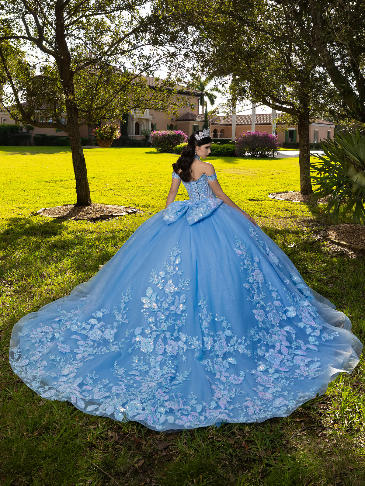 A woman stands outdoors on a grassy lawn wearing a voluminous light blue ball gown with floral applique and a matching blue sash, photographed from behind in a park-like setting with trees and buildings in the background. / This ball gown features an embellished bodice, off-the-shoulder sleeves, and a full skirt covered in detailed floral appliqué. The back is finished with a large decorative bow.