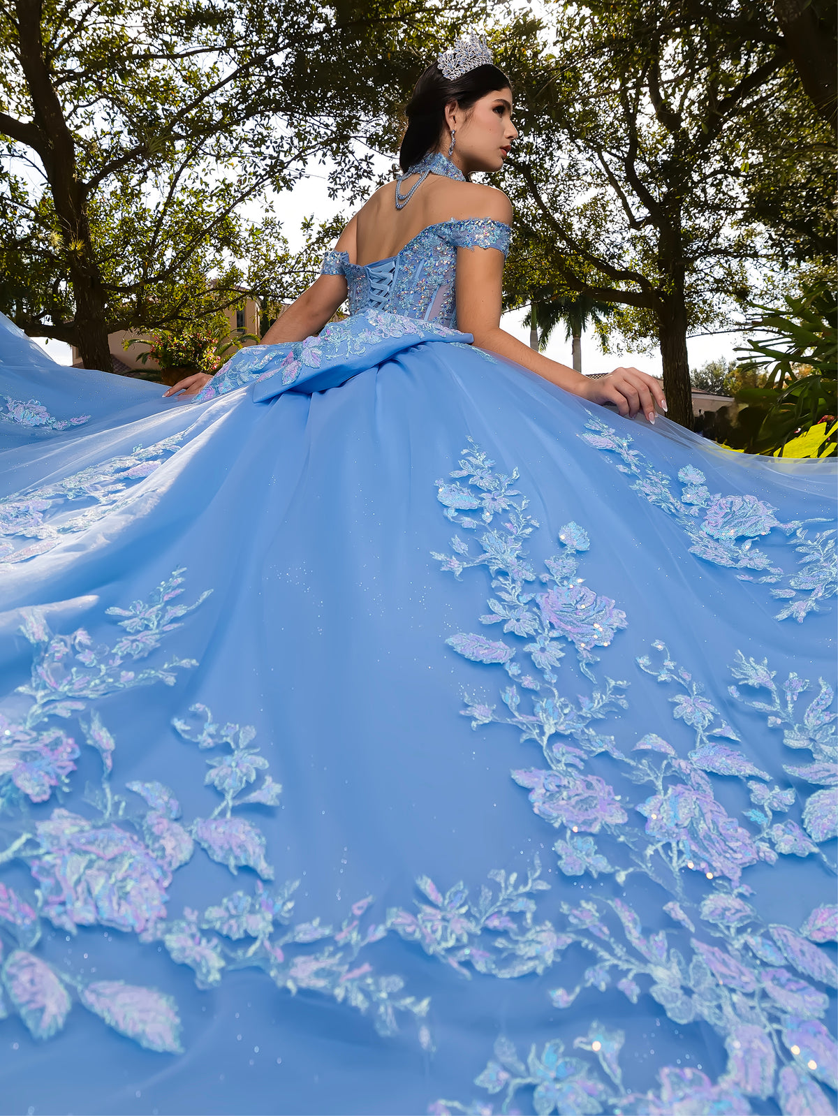 A woman wearing a voluminous light blue quinceañera ball gown with floral embroidery, photographed outdoors with trees in the background. She is seen from behind, showcasing the back of the dress and its large train. / This ball gown features an embellished bodice, off-the-shoulder sleeves, and a full skirt covered in detailed floral appliqué. The back is finished with a large decorative bow.