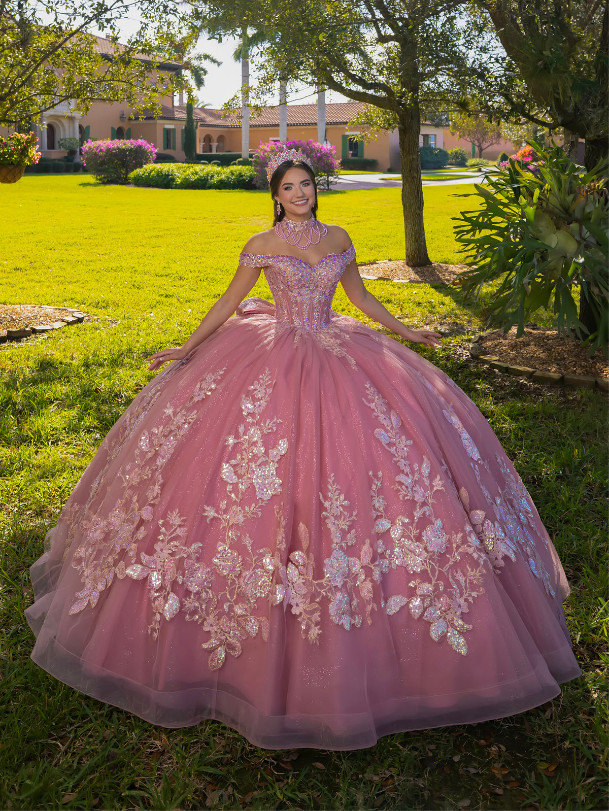 A young woman wearing a voluminous dusty pink ball gown with floral lace appliqués, standing on a green lawn in front of a large house and trees. / This ball gown features an embellished bodice, off-the-shoulder sleeves, and a full skirt covered in detailed floral appliqué. The back is finished with a large decorative bow.