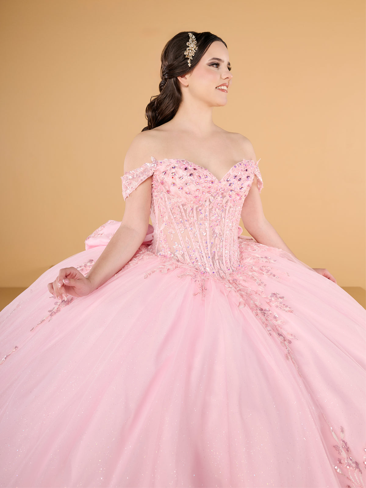 A woman modeling a voluminous light pink off-the-shoulder ball gown with a heavily embellished corset bodice and a full tulle skirt, posed against a solid warm beige background. / This ball gown features an embellished off-the-shoulder bodice and a full skirt with detailed floral accents.