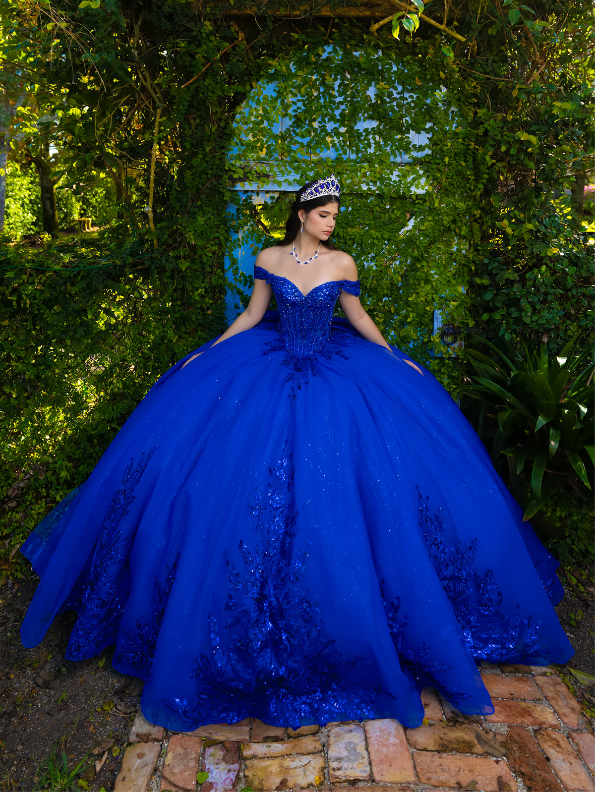 A woman wearing a voluminous royal blue off-the-shoulder ball gown with lace applique and a tiara, standing outdoors in front of a leafy green arched doorway. / This ball gown features an embellished off-the-shoulder bodice and a full skirt with detailed floral accents.