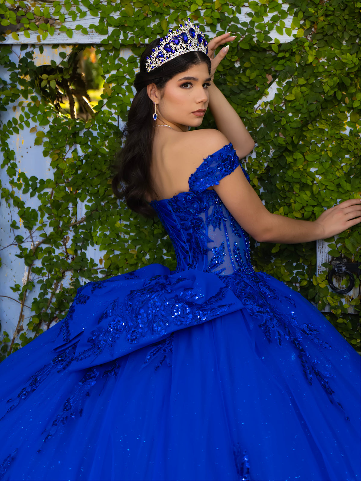 A woman wearing a royal blue off-the-shoulder ball gown with sequined embroidery and a matching tiara, posing outdoors against a wall covered in green ivy. / This ball gown features an embellished off-the-shoulder bodice and a full skirt with detailed floral accents.
