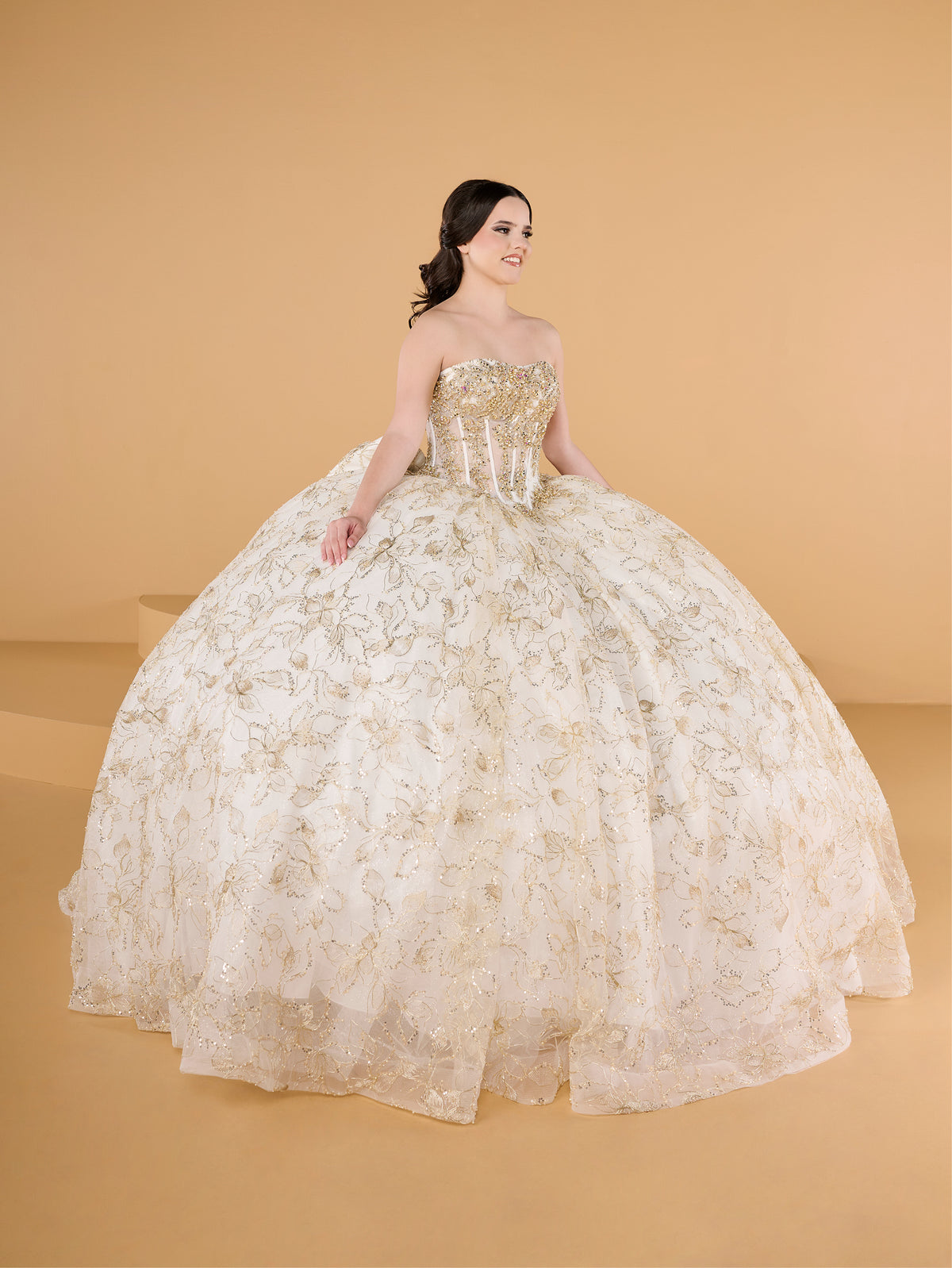 A woman modeling a strapless ball gown with a voluminous tulle skirt and gold floral embroidery, set against a solid warm beige studio background. / This ball gown features an embellished off-the-shoulder bodice, and a full skirt accented with detailed floral designs, finished with matching appliqué on the back.