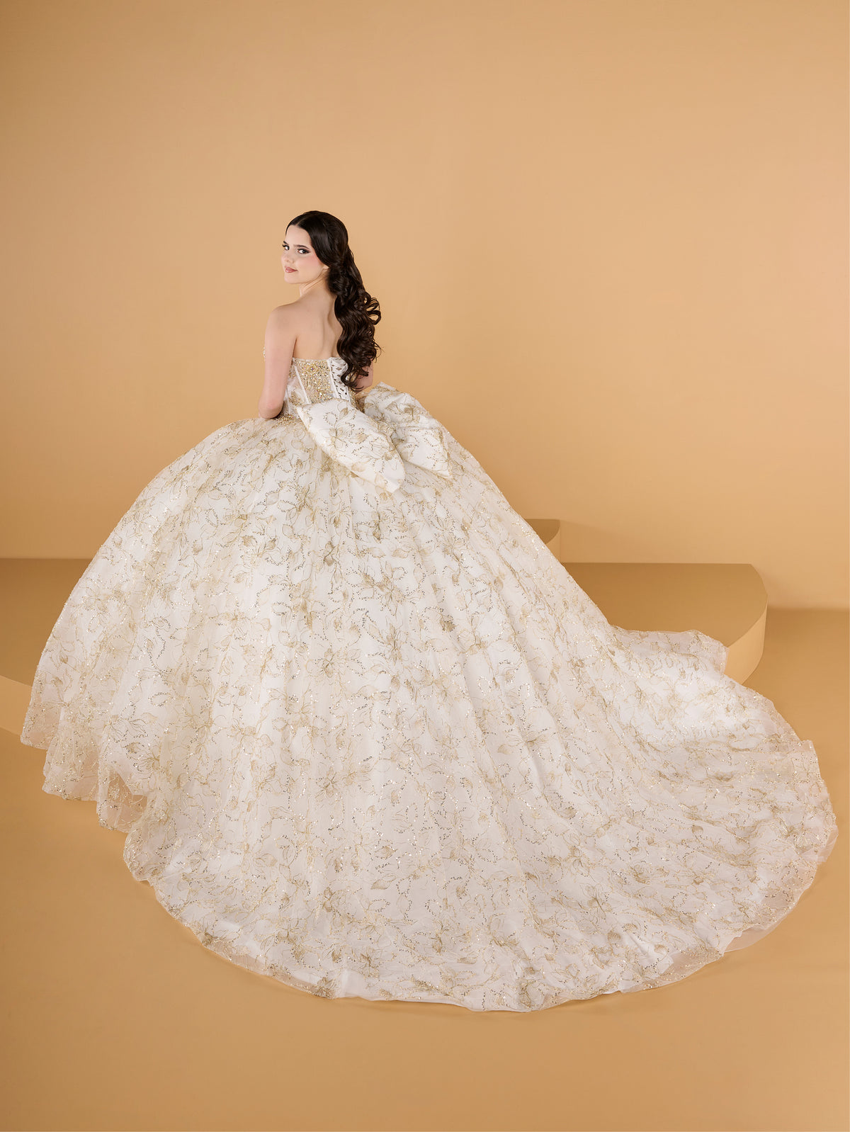 A woman modeling a strapless ball gown with a voluminous floral lace skirt and long draped sleeves, photographed against a warm beige studio backdrop. / This ball gown features an embellished off-the-shoulder bodice, and a full skirt accented with detailed floral designs, finished with matching appliqué on the back.