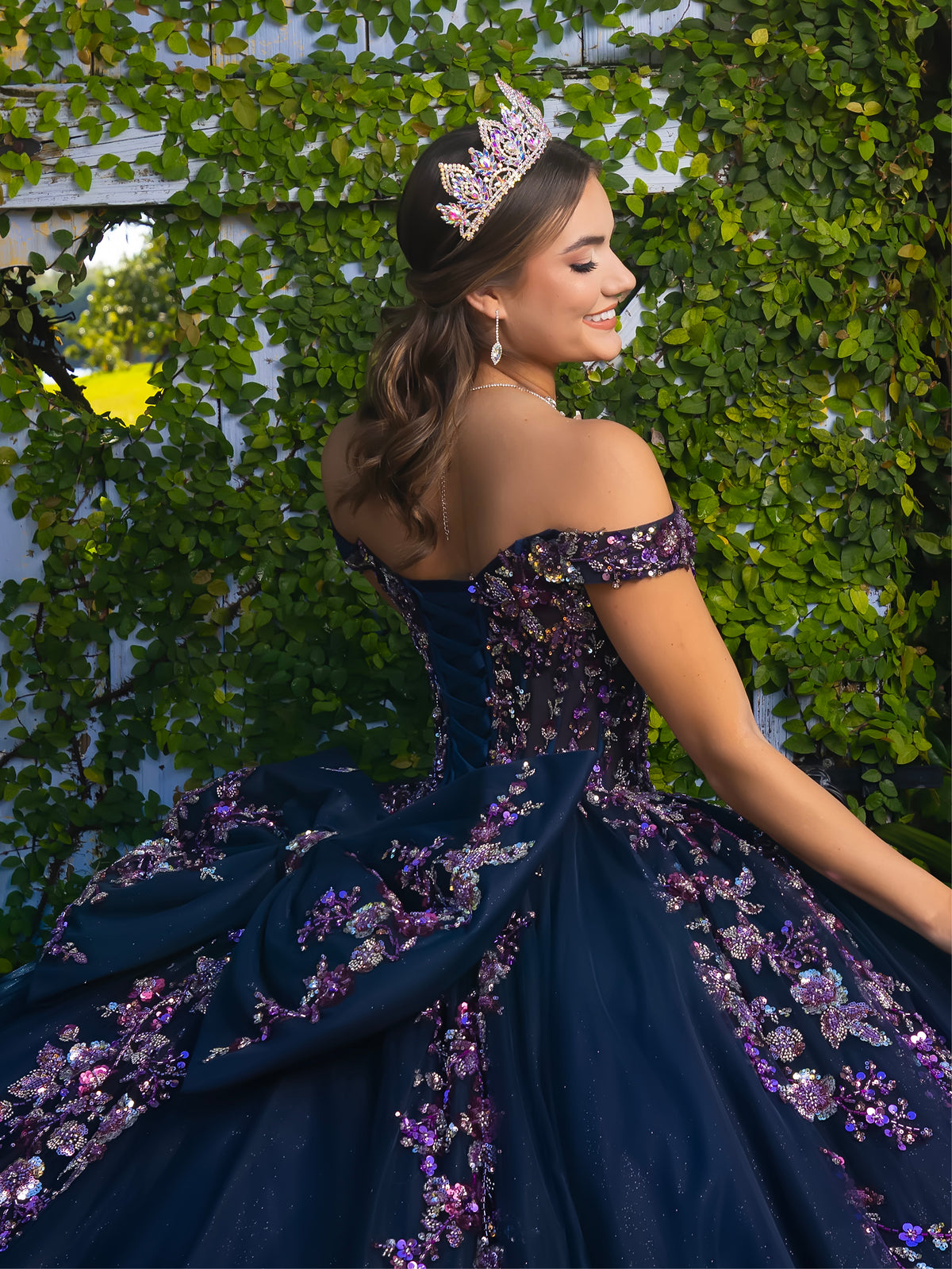 A woman wearing a dark navy off-the-shoulder ball gown with purple and pink floral sequin embellishments, posing outdoors against a green ivy-covered wall while wearing a jeweled tiara. / This ball gown features an embellished bodice with off-the-shoulder sleeves and a full tulle skirt adorned with delicate floral appliqué. A large bow accents the back.