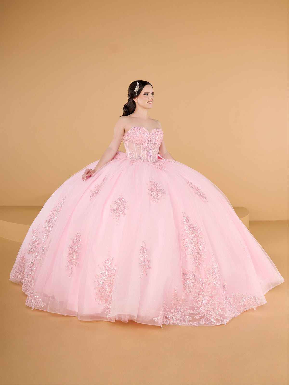 A woman modeling a voluminous light pink ball gown with a strapless sweetheart bodice and embroidered floral details, set against a solid warm beige studio background. / This ball gown features a fitted, embellished bodice and a full skirt decorated with floral appliqué, finished with a bow at the back.
