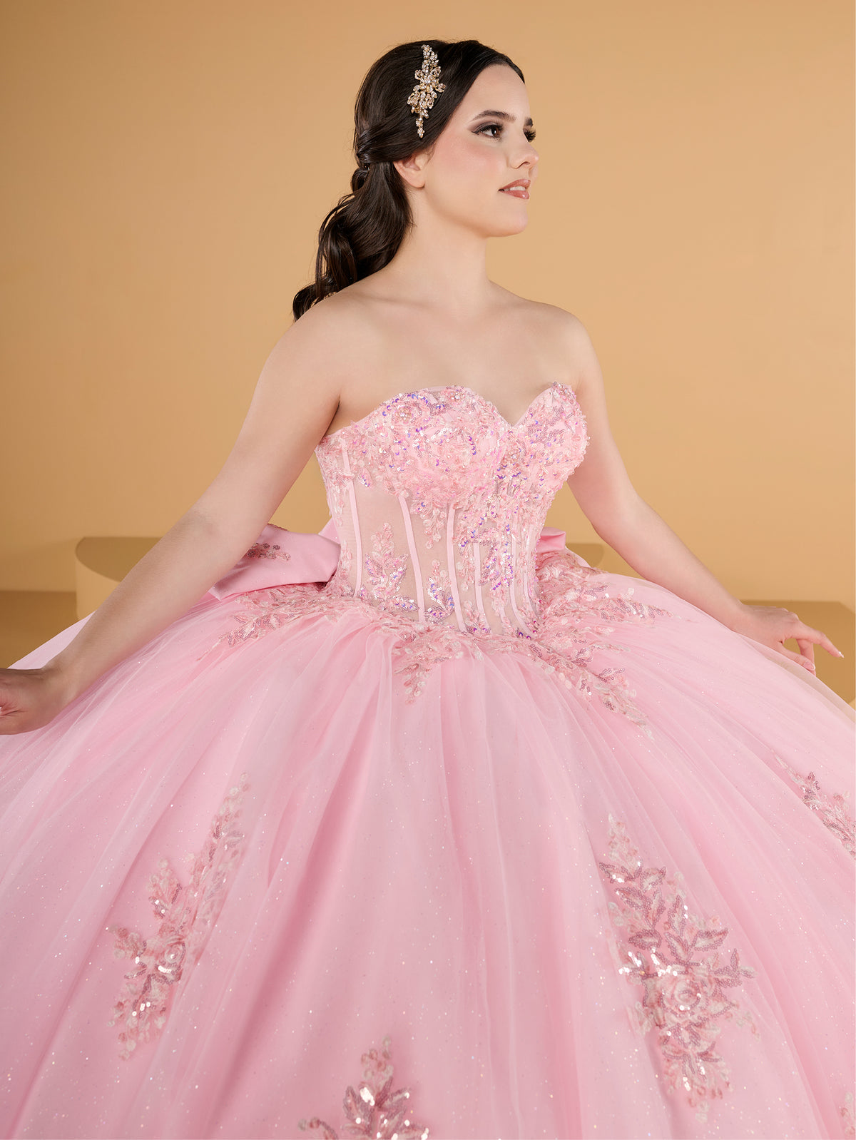 Woman wearing a pink quinceañera dress with floral embellishments against a yellow background / This ball gown features a fitted, embellished bodice and a full skirt decorated with floral appliqué, finished with a bow at the back.