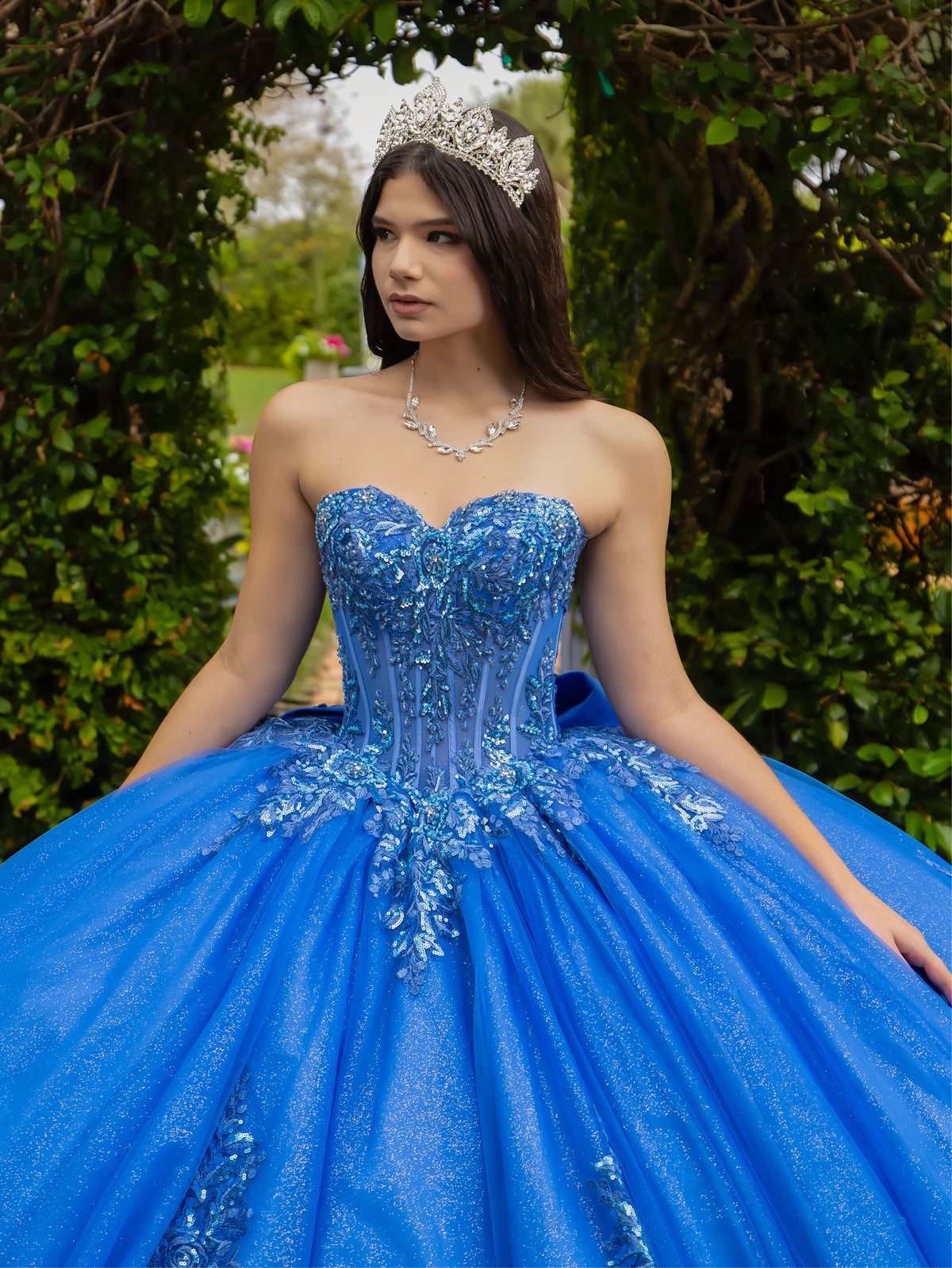 Woman in a blue quinceañera dress with a tiara against a green outdoor background / This ball gown features a fitted, embellished bodice and a full skirt decorated with floral appliqué, finished with a bow at the back.