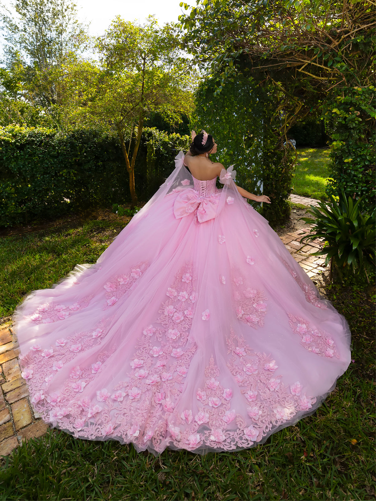 A young girl viewed from behind wearing a voluminous light pink ball gown with 3D floral appliqués and a large bow on the back, standing in a lush green garden. / This ball gown features a floral-embellished bodice and a full tulle skirt with cascading 3D flowers, finished with a decorative bow at the back.