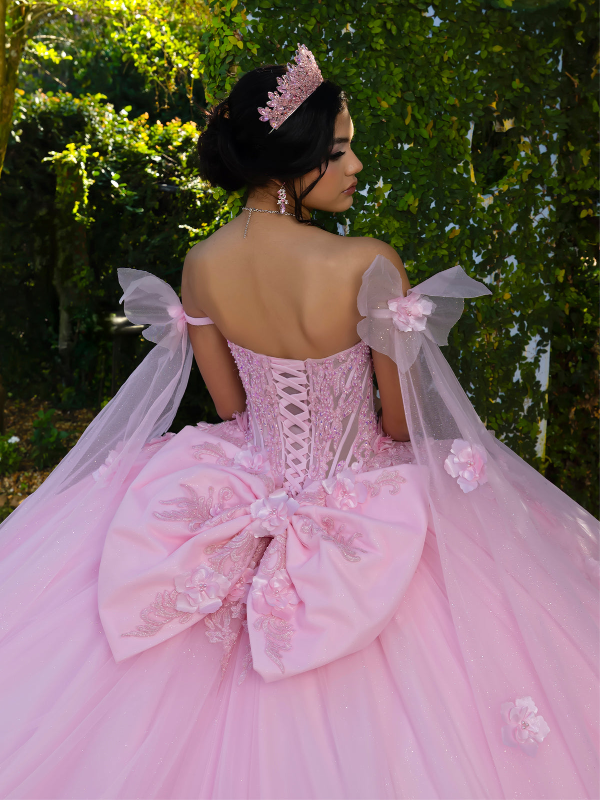 Woman in a pink dress with floral details and a tiara, standing against a green leafy background. / This ball gown features a floral-embellished bodice and a full tulle skirt with cascading 3D flowers, finished with a decorative bow at the back.