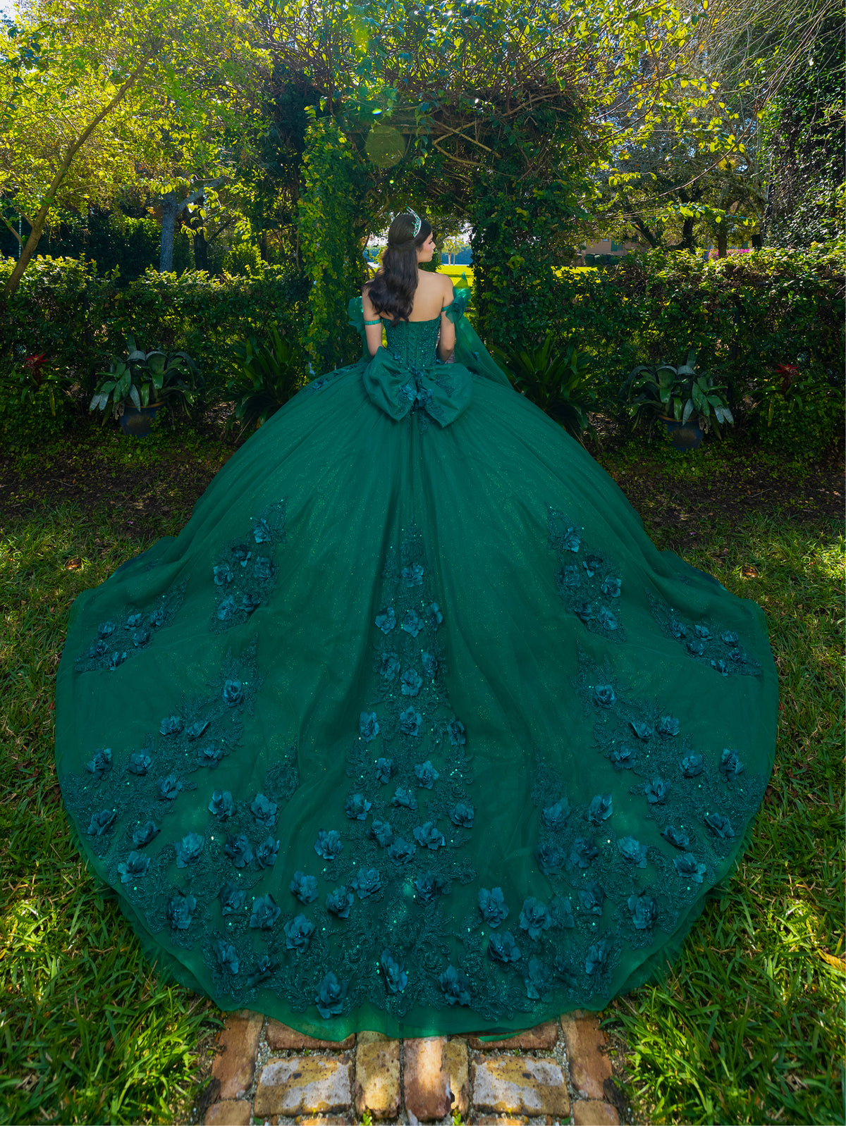 A woman stands outdoors in a lush garden wearing a dramatic emerald green ball gown from behind, showing an enormous train with 3D floral appliqués and a large bow at the back of the neckline. / This ball gown features a floral-embellished bodice and a full tulle skirt with cascading 3D flowers, finished with a decorative bow at the back.
