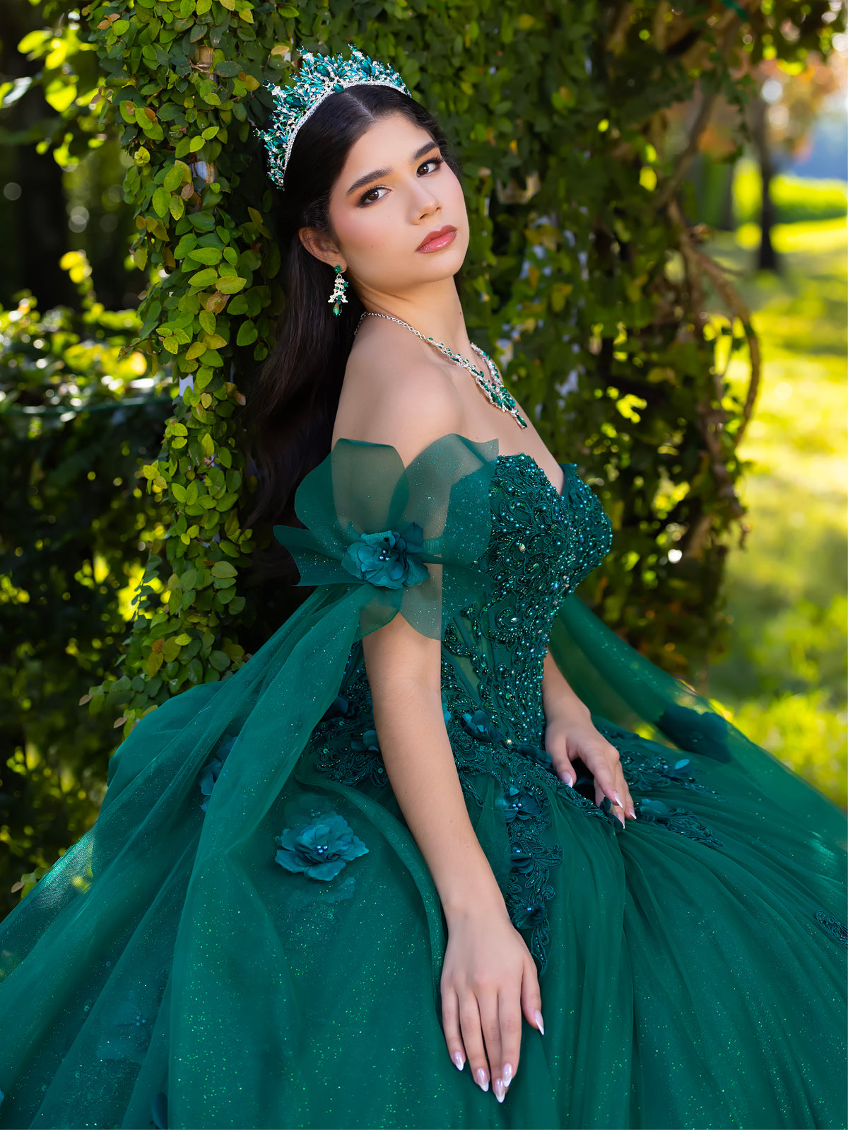 A young woman wearing a voluminous emerald green quinceañera ball gown with off-the-shoulder sheer fabric, beaded bodice, and matching green tiara and earrings, posed outdoors against a leafy green background. / This ball gown features a floral-embellished bodice and a full tulle skirt with cascading 3D flowers, finished with a decorative bow at the back.