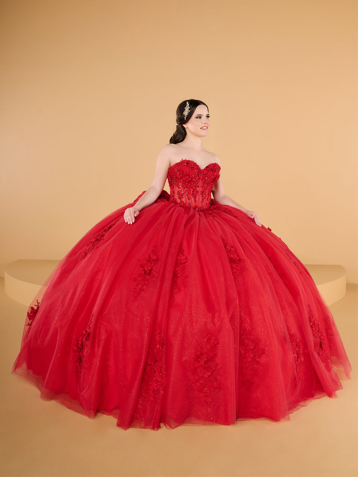 A woman modeling a voluminous red ball gown with a strapless sweetheart bodice, floral lace appliqués, and a full tulle skirt against a warm beige studio backdrop. / This ball gown features a fitted, embellished bodice with thin straps and a full skirt decorated with floral appliqué, finished with a bow at the back.