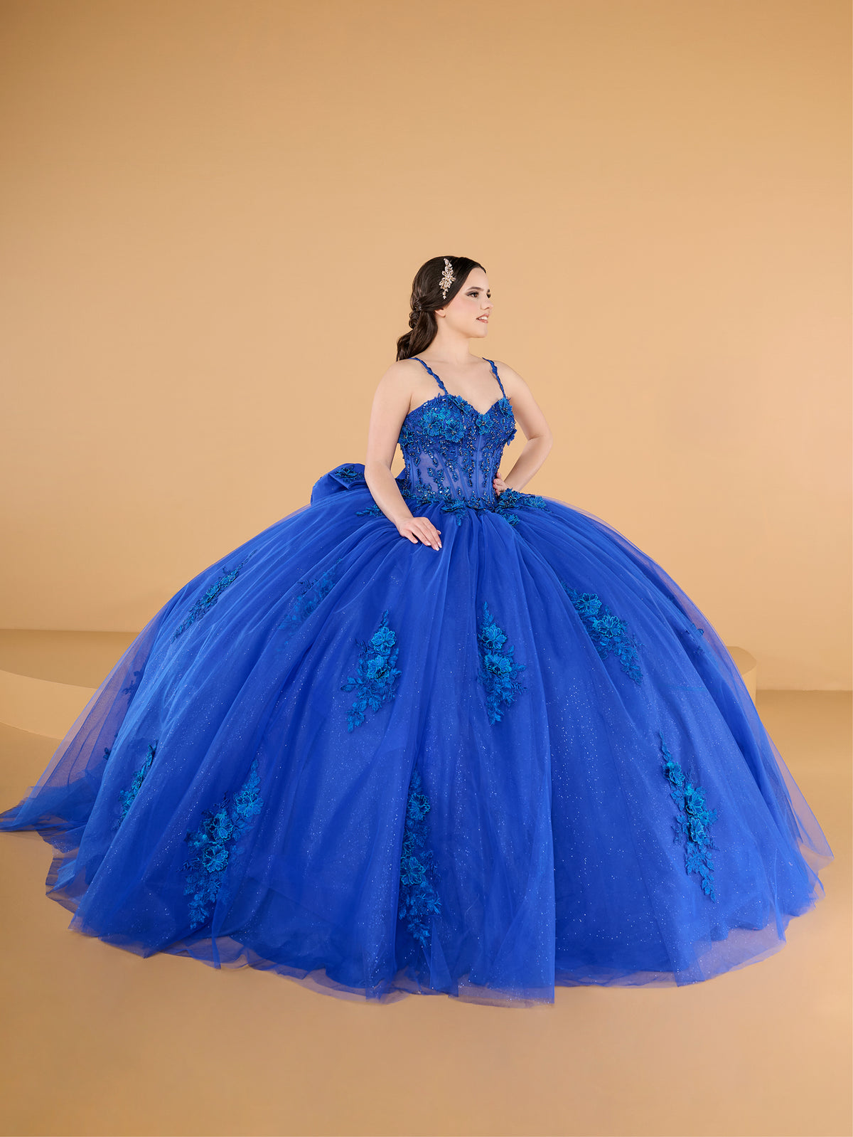 A woman modeling a voluminous royal blue ball gown with floral lace applique and a sweetheart neckline, posed against a solid beige studio backdrop. / This ball gown features a fitted, embellished bodice with thin straps and a full skirt decorated with floral appliqué, finished with a bow at the back.