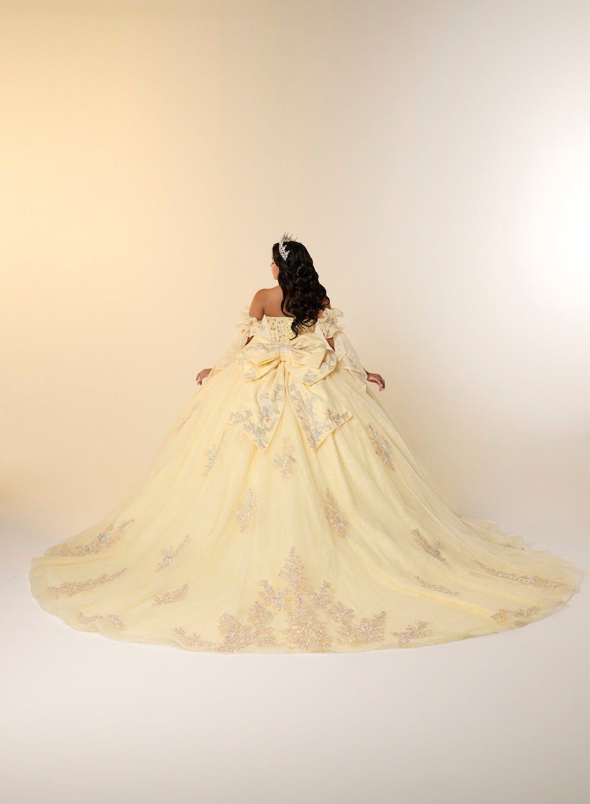 A woman viewed from behind wearing an off-the-shoulder ball gown with a very full skirt and long train in pale yellow with silver floral embroidery, set against a warm beige studio background. / This elegant ball gown features a strapless straight neckline with delicate draped sleeves. The full skirt is crafted from soft tulle and adorned with shimmering embroidery, while a large bow at the back adds a graceful finishing touch.