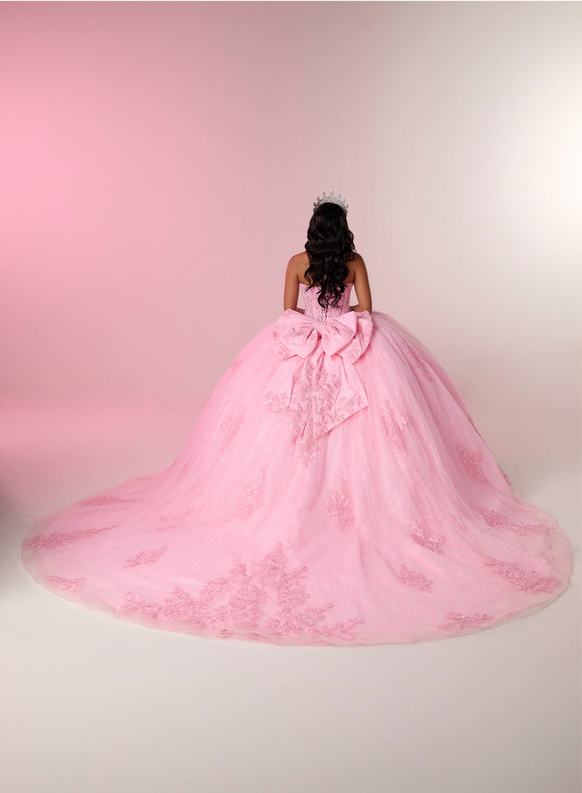 A woman viewed from behind wearing a voluminous light pink ball gown with a large bow at the back, set against a soft pink and white studio backdrop. / This elegant ball gown features a strapless straight neckline with delicate draped sleeves. The full skirt is crafted from soft tulle and adorned with shimmering embroidery, while a large bow at the back adds a graceful finishing touch.