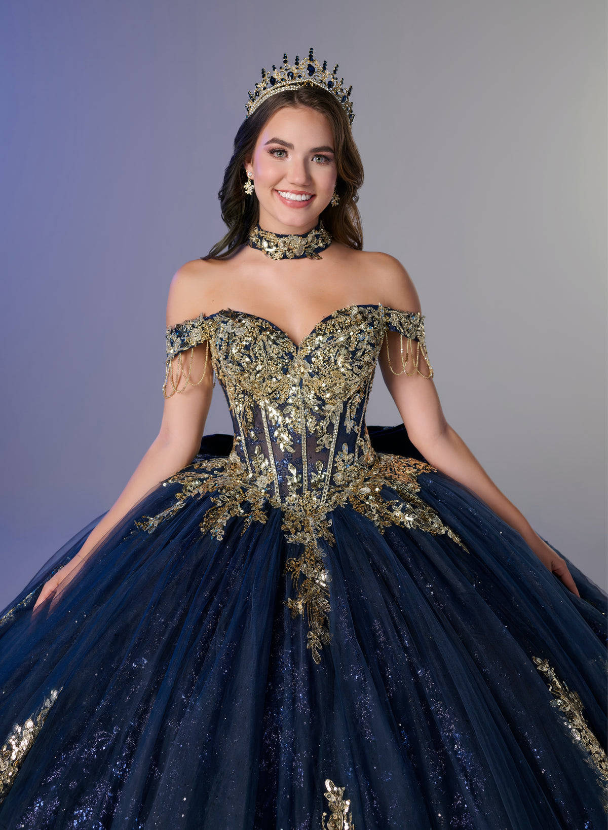 A woman modeling an elaborate off-the-shoulder ball gown in deep navy blue with extensive gold sequin and bead embroidery, accessorized with a matching gold choker and crown, set against a solid gray studio background. / An off-the-shoulder ball gown featuring an embroidered bodice, a voluminous tulle skirt with floral applique panels, and a large accent bow on the back.