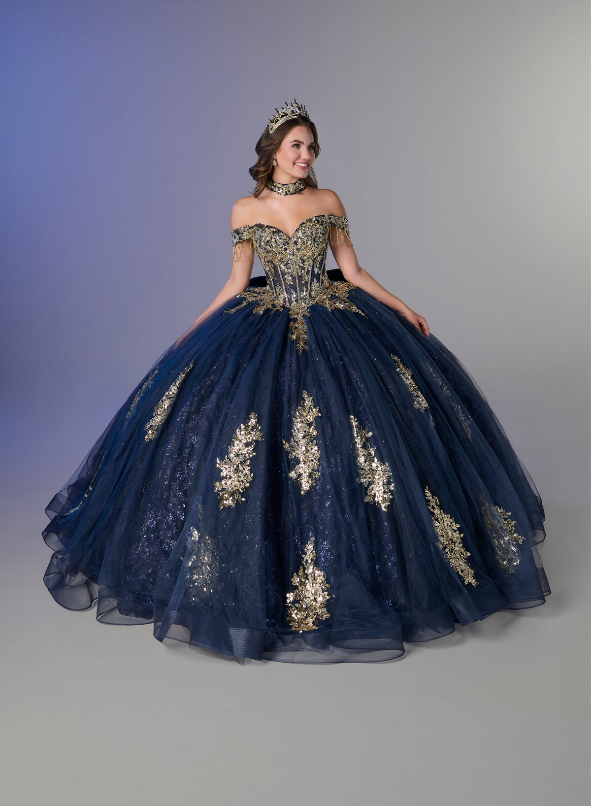 A young woman modeling an off-the-shoulder navy blue ball gown with intricate gold embroidery and a matching tiara, set against a gray studio background. / An off-the-shoulder ball gown featuring an embroidered bodice, a voluminous tulle skirt with floral applique panels, and a large accent bow on the back.